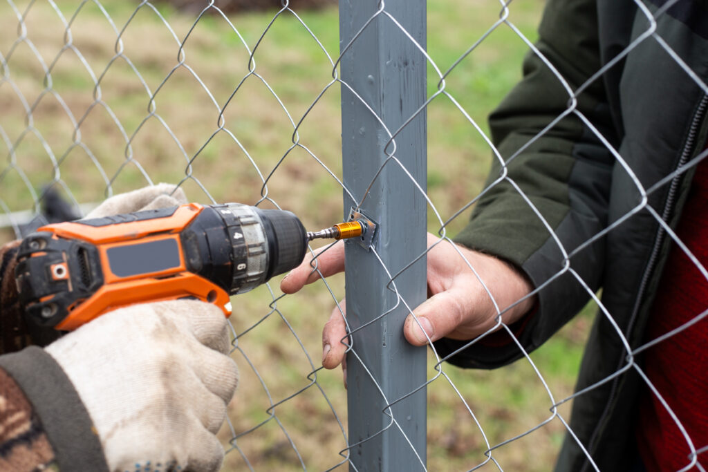 A man is building a fence at his dacha. Using an electric drill, he screws a metal mesh chain-link to the metal profile.